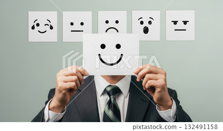 A person in a suit holds a card with a happy emoji, with other facial expressions in the background, symbolizing satisfaction. 132491158