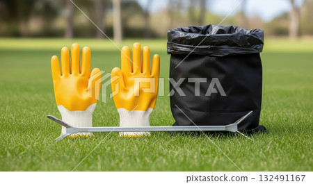 Yellow gardening gloves and a black trash bag sit on lush green grass, accompanied by a metal litter picker, promoting eco-friendly waste management practices in outdoor spaces 132491167
