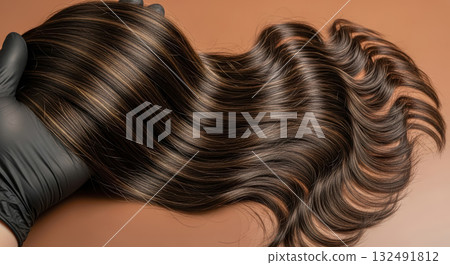 Close-up of long, shiny, wavy brown hair with highlights, held by a hand in a black glove 132491812