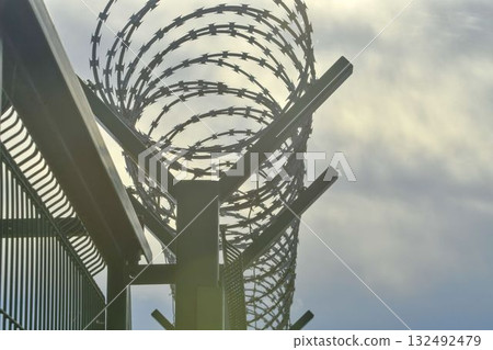 Barbed razor wire on a high-security building. Barbed wire and hard wire fence. Concepts for jails, army facilities, nuclear power plants, and defense against illegal migration. Barbed razor wire on a high-security building. Barbed wire and hard wire fence. Concepts for jails, army facilities, nuclear power plants, and defense against illegal migration. 132492479