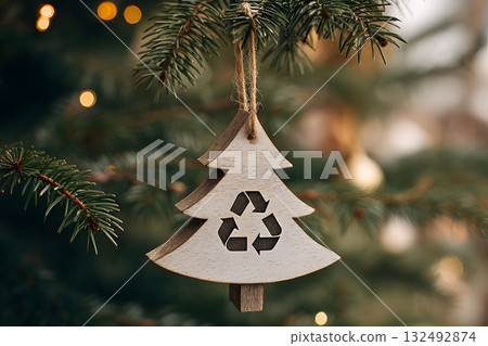 A wooden Christmas tree ornament with a recycling symbol hangs from a pine branch. The background features soft, warm lights, emphasizing sustainable holiday decor. 132492874