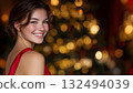 Smiling young Caucasian woman with brown hair in a red dress, standing in front of a decorated Christmas tree with warm lights, celebrating New Year 2026. 132494039
