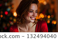 Smiling young Caucasian woman with wavy brown hair, wearing a red dress, in front of a decorated Christmas tree with warm lights, celebrating New Year 2026. 132494040