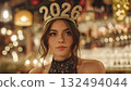 A young Caucasian woman with brown hair wears a sparkling 2026 headband. She is in a festive setting with blurred lights and decorations, celebrating New Year's Eve. 132494044