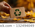 A hand holds a wooden card with a green recycling symbol. The background features autumn leaves, emphasizing themes of ecology and sustainability. 132494049