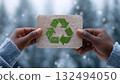 A young Black woman holds a recycled paper card with a green recycling symbol. Snowy forest background emphasizes themes of ecology and sustainability. 132494050