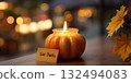 A lit pumpkin candle with a 'Give Thanks' card on a wooden table. Autumn leaves are visible in the background, creating a warm, festive atmosphere for Thanksgiving. 132494083