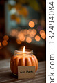 A pumpkin-shaped candle with a lit flame sits on a wooden table. A card with the words 'Give Thanks' is placed beside it. Soft bokeh lights create a warm autumn atmosphere. 132494084