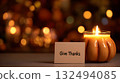 A lit pumpkin-shaped candle beside a card that reads 'Give Thanks'. The background features warm, blurred autumn lights, creating a cozy Thanksgiving atmosphere. 132494085