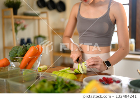 Cropped shot woman slicing a green apple in her bright home kitchen, surrounded by fresh vegetables and fruits 132494241