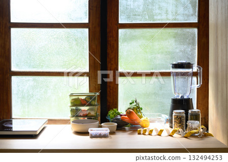 Healthy lifestyle setup with vegetables, smoothie blender, food storage containers, and measuring tape on a wooden kitchen counter by the window, symbolizing wellness and nutrition planning 132494253