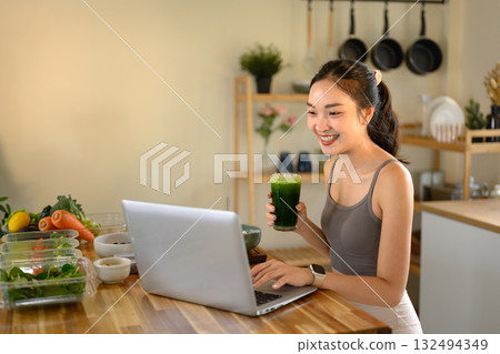 Smiling young woman drinking a green smoothie while working on a laptop in her cozy kitchen 132494349