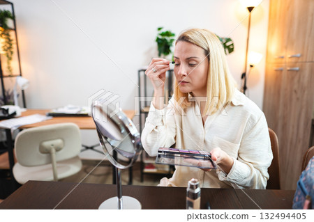 Woman applying makeup in a cozy home office with warm lighting 132494405