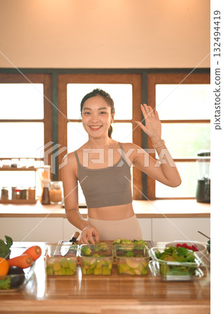 A smiling young woman waving while preparing healthy vegetable meals in her kitchen, expressing friendliness and positive energy in a healthy lifestyle A smiling young woman waving while preparing healthy vegetable meals in her kitchen, expressing friendliness and positive energy in a healthy lifestyle 132494419