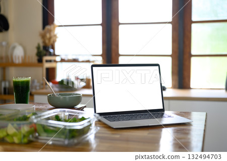 Laptop with blank white screen on a wooden kitchen counter beside a green smoothie and healthy food 132494703