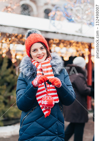 Young woman on christmas market. cute lovely girl in red hat standing at street and hangs ball on Christmas tree outdoors in winter snow day. Christmas lights 132498007