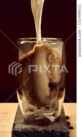 Milk pouring into glass of dark coffee with swirling texture and roasted beans on stone block Milk pouring into glass of dark coffee with swirling texture and roasted beans on stone block 132499011