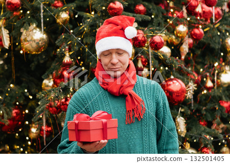 Portrait of happy man wearing green sweater and Santa Hat holding red gift box near Christmas tree. Xmas Holiday. Close up. 132501405