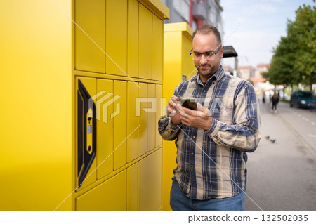 Man using smartphone while standing near bright yellow delivery lockers in an urban environment during daytime 132502035
