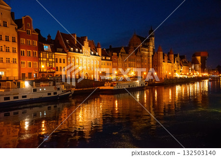 Gdansk riverside at night showcases historic buildings beautifully lit, reflecting on Motlawa River. People stroll along the promenade, enjoying a peaceful evening atmosphere 132503140