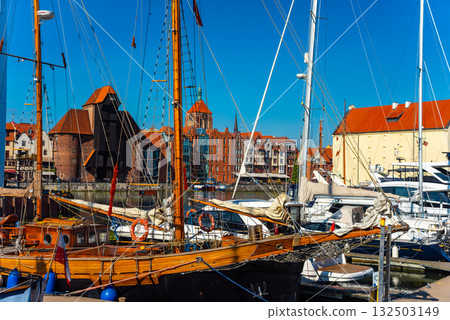 A traditional wooden boat is anchored at the Gdansk marina against a backdrop of vibrant, historical architecture. The clear blue sky enhances the picturesque scene. 132503149