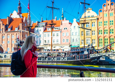 Tourist in red jacket with backpack walks along Gdansk waterfront. Behind her are historical buildings with vibrant facades and a traditional sailing ship at the harbor. 132503151