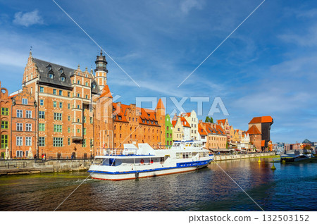 Scenic view of two boats navigating the Motlawa River alongside historic colorful buildings in Gdansk, Poland. Bright blue skies enhance this vibrant European cityscape 132503152