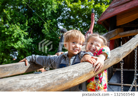 Two cheerful children enjoy sunny day at wooden playground surrounded by green trees. Laughter and bright colors create joyful atmosphere of friendship and childhood happiness Two cheerful children enjoy sunny day at wooden playground surrounded by green trees. Laughter and bright colors create joyful atmosphere of friendship and childhood happiness 132503540