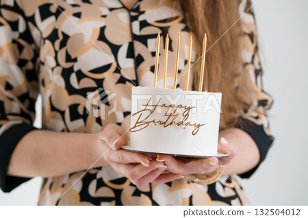 Hands Holding Small White Birthday Cake with Gold Candles and Happy Birthday Text, Creamy Frosting, Modern Minimalist Style. 132504012