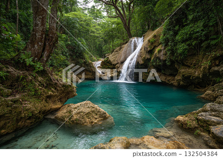A stunning jungle waterfall cascades into a vibrant turquoise pool surrounded by lush green foliage and large brown rocks, creating a secluded natural oasis. 132504384