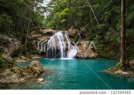 A stunning jungle waterfall cascades into a vibrant turquoise pool surrounded by lush green foliage and large brown rocks, creating a secluded natural oasis. 132504385