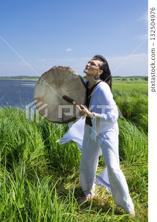 Middle-aged female shaman in the white dress drumming outdoors Middle-aged female shaman in the white dress drumming outdoors 132504976