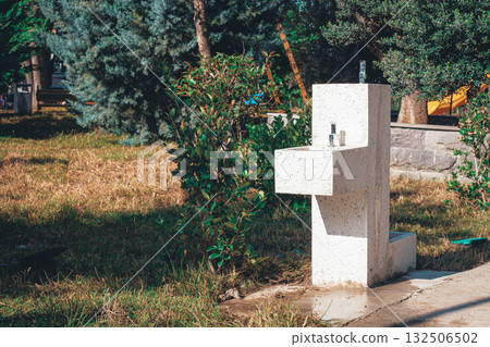 Water fountain stands in a sunny park surrounded by greenery and grass near a walking path 132506502