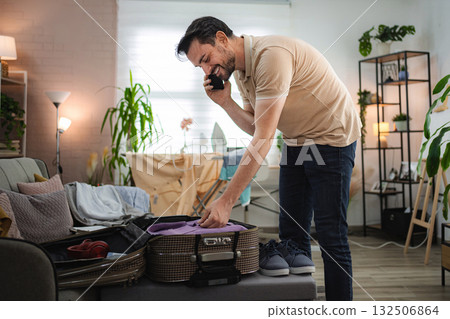Man talking on the phone while packing suitcase at home 132506864