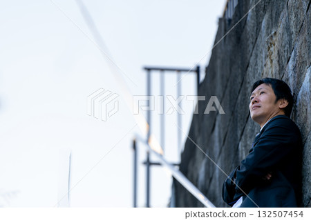 Businessman leaning against a stone wall and looking to the future 132507454