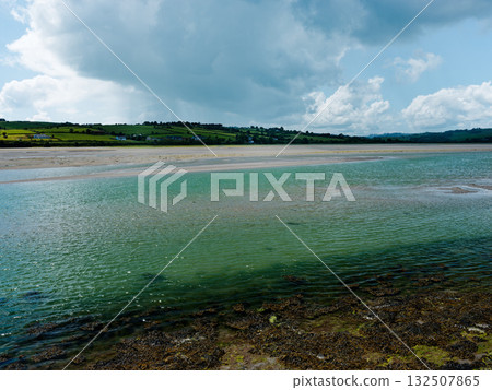 Tranquil scenery of a bay where clear, green-tinted water merges with a sandy bank. Rolling hills dotted with houses sit calmly in the background under a bright skyline. 132507865