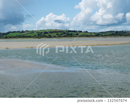 Scenic view of calm waters and a sandy beach, transitioning to green hills under a wide open sky and scattered white clouds. Scenic view of calm waters and a sandy beach, transitioning to green hills under a wide open sky and scattered white clouds. 132507874