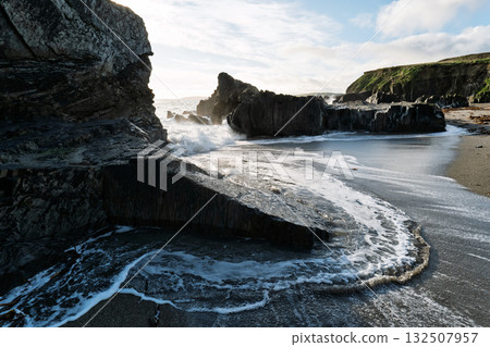 Waves from the ocean gently wash up on the black sand beach among the rocks and cliffs at Sheep Cove in Ireland on a bright day with fluffy clouds. 132507957