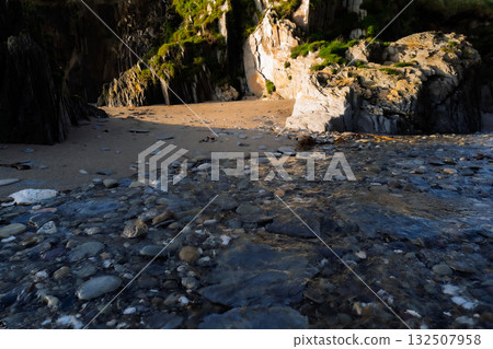 Water rushes over rocks at low tide as it flows toward a sandy cove surrounded by rocky cliffs. The scene is captured in West Cork, Ireland. 132507958