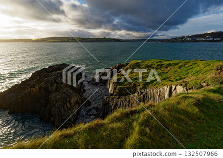 Overlooking the ocean from a grassy cliff in West Cork, Ireland. Waves move around the rock formations. The sun shines through the cloudy sky, creating a scenic landscape. 132507966