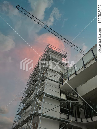 Scaffolding is seen surrounding a tall building under construction as dawn breaks. A crane towers above, contrasting the pastel colors of the morning sky. 132508032
