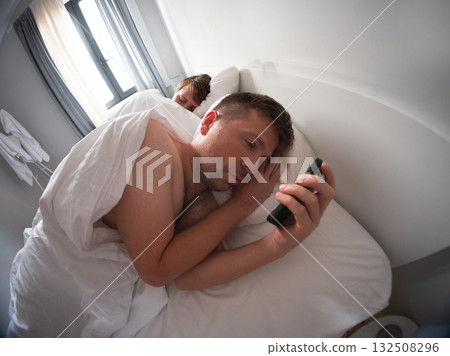 Man using his phone at night while his wife sleeps beside him, bad habit and screen addiction Man using his phone at night while his wife sleeps beside him, bad habit and screen addiction 132508296