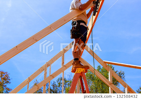 Construction professional carefully positions roof truss, balancing on ladder 132508432