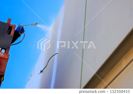 Worker uses spray tool apply paint on building wall while elevated on lifting equipment Worker uses spray tool apply paint on building wall while elevated on lifting equipment 132508433