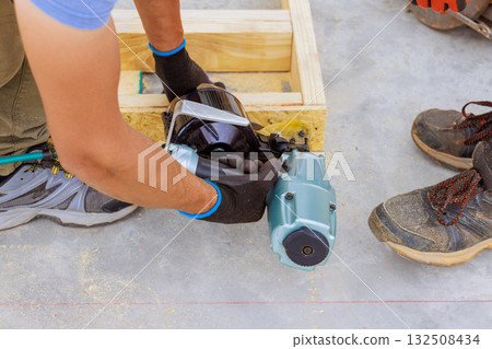 Worker is assembling wooden frame using power air nail gun while standing on construction site. 132508434