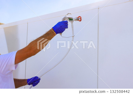 Worker with gloves applies paint to white wall using paint sprayer gun Worker with gloves applies paint to white wall using paint sprayer gun 132508442