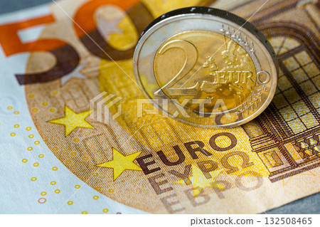 Two euro coin standing on fifty euro banknote, symbolizing European finance, savings, and economy Two euro coin standing on fifty euro banknote, symbolizing European finance, savings, and economy 132508465