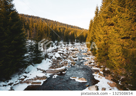 A serene winter evening at the Jizera River, marking the border between Czechia and Poland. Snow covers the ground, and tall evergreen trees line the riverbank, creating a peaceful scene. A serene winter evening at the Jizera River, marking the border between Czechia and Poland. Snow covers the ground, and tall evergreen trees line the riverbank, creating a peaceful scene. 132508654