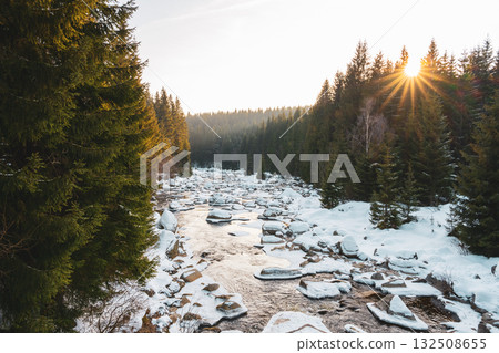 Snow-covered banks line the calm Jizera River during a winter evening. Sunlight shines through evergreen trees, creating a peaceful atmosphere at the Czechia and Poland border. 132508655