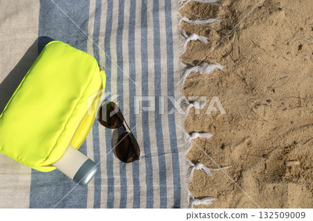Flat lay with a makeup bag, SPF cream, and dark sunglasses on a beach towel at the beach. 132509009